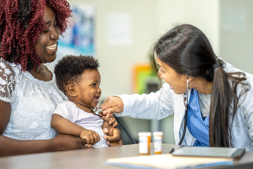A cute baby boy sits on his mother's lap and is seen by a young female doctor. There are pill bottles on the table and the mother and doctor are discussing treatment options.