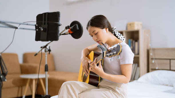 Asian woman playing guitar and recording vocals in her bedroom. She's live. stock photo