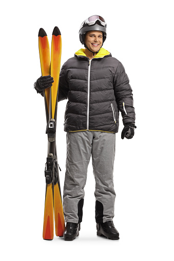 Full length portrait of a man in a skiing jacket and boots holding pair of skis isolated on a white background