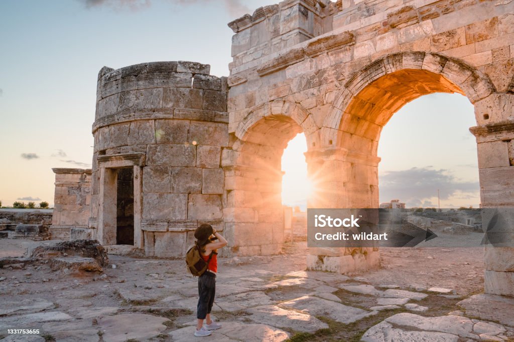 Photographer tourist girl is taking photos of the Frontinus Gate in ancient ruins in Hierapolis , Pamukkale UNESCO, Backpacker, Camera, Travertine pools, Greek architecture Türkiye - Country Stock Photo