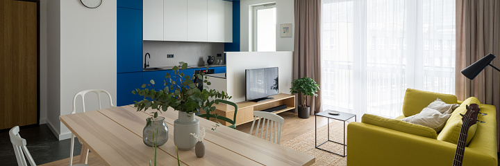 Panorama of long, wooden dining table with different chairs in modern apartment with small, blue kitchen open to living room with yellow couch and tv