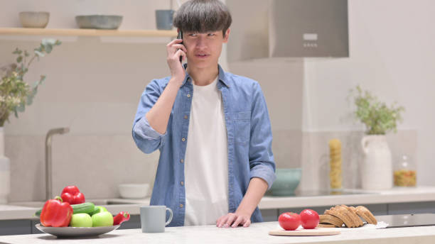 istock Young Asian Man Talking on Smartphone while Standing  in Kitchen 1331795129