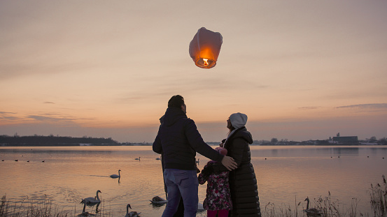Family in warm clothing releasing burning sky lantern and admiring from riverbank