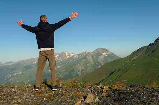 Hiker standing with arms outstretched up to the sky. Snow-capped mountain peaks in the background.