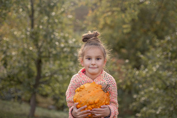 Happy smilling child girl in a knitted sweater is holding in hands ugly orange pumpkin. Deformed orange pumpkin with a damaged, ugly skin. Thanksgiving, harvest, halloween concept.
