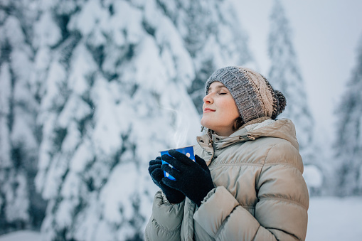 Young woman holding coffee cup and standing on snow covered forest with eyes closed