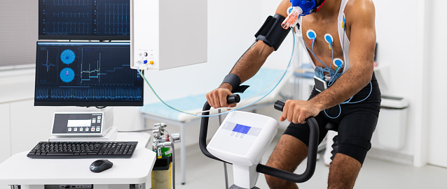 Doctors observing the progress of a cardiopulmonary stress test taken by the male athlete riding a bicycle ergometer. Young adult man having a VO2 test with a VO2 mask on his face, electrocardiogram pads attached, computer recording, indoor bicycle. Healthcare and Medicine concept.