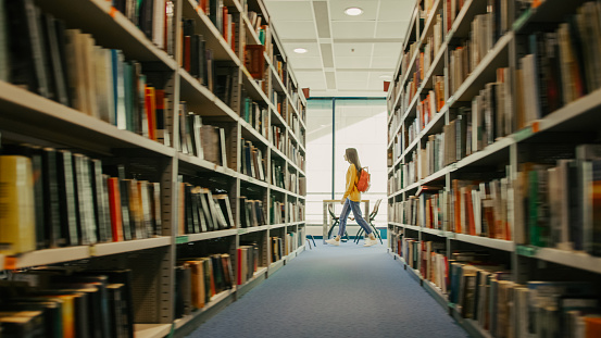 Female student walking beside bookshelves in library