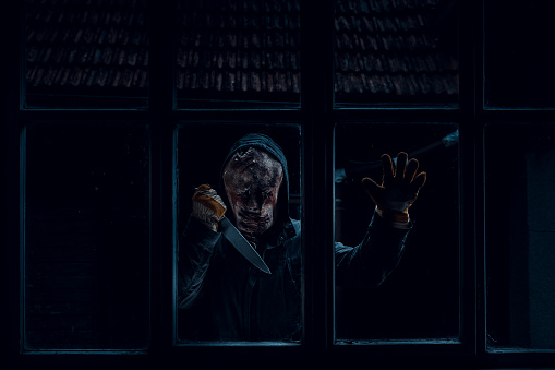 Creepy man holding a big sharp knife, looking at the camera, standing outside the window in the dark. Scene from horror, man in disguise standing outside the window in the dark.