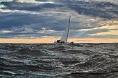 The lonely sailboat on the horizon in sea at sunset, the storm sky of different colors, big waves, sail regatta, cloudy weather, only main sail, sun beams