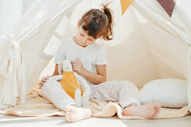 Six years cute girl playing in teepee with fabric tilda bunny at home
