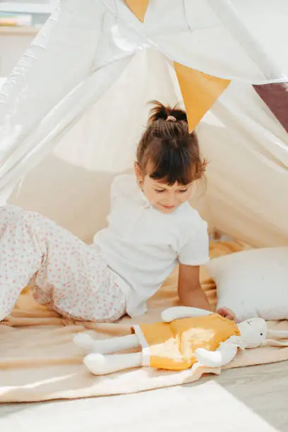 Six years cute girl playing in teepee with fabric tilda bunny at home