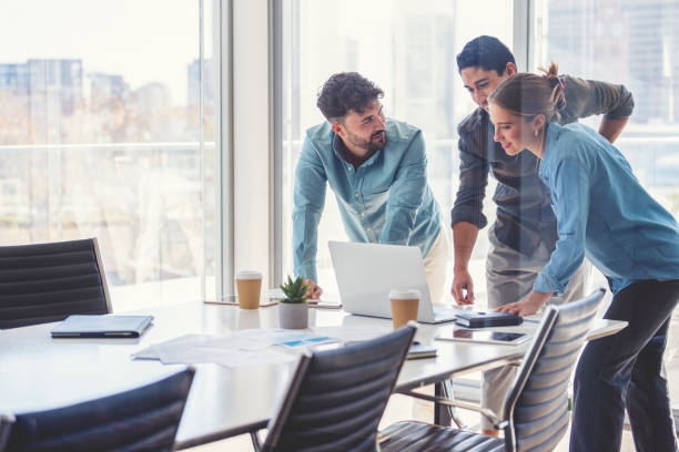 Business team working on a laptop computer. Business team working on a laptop computer. Three people are wearing casual clothing. They are standing in a board room. Multi ethnic group with Caucasian and Latino men and women. They are all happy and smiling office window night stock pictures, royalty-free photos & images