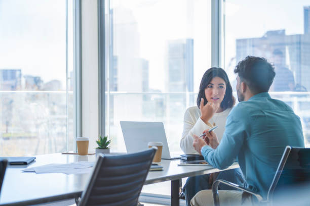 Business woman and man meeting and talking Business woman and man meeting and talking. Both are casually dressed. There is a laptop computer on the table. They are sitting at a boardroom table with a window behind them business stock pictures, royalty-free photos & images