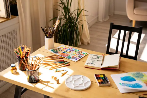 Photo of The artist's workplace with watercolor paints, gouache, brushes and sheets of paper on a wooden background. Copy space. Flat lay, top view