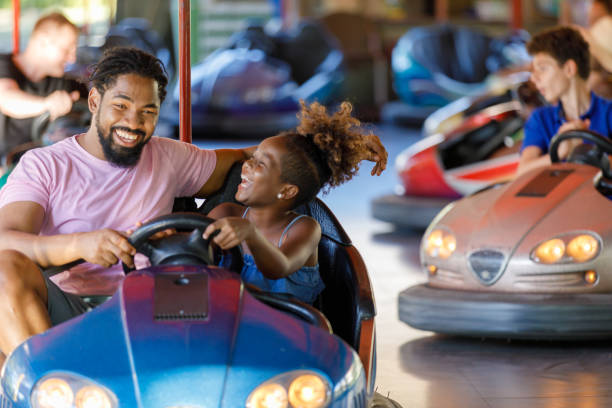 African-American Father is Driving in Bumper Car with his Cute Little Daughter. A Beautiful Little Daughter is Happy to Spend a Wonderful Day with her Happy Father. They are Driving an Electric Car in the Amusement Park. amusement park stock pictures, royalty-free photos & images