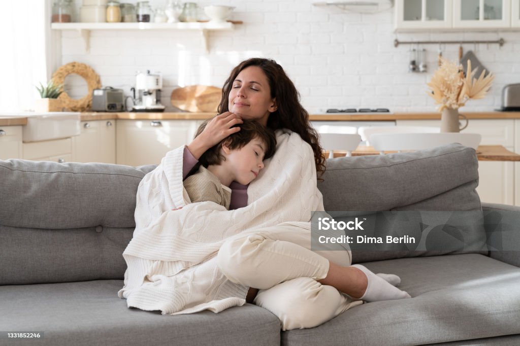 Young mom sit on cozy couch relax with preschool son covered with blanket on weekend morning at home Young mom sit on cozy couch relax with preschool child covered with blanket bonding with kid on weekend morning at home. Loving mother single parent wrapping embrace small son on sofa in living room Blanket Stock Photo