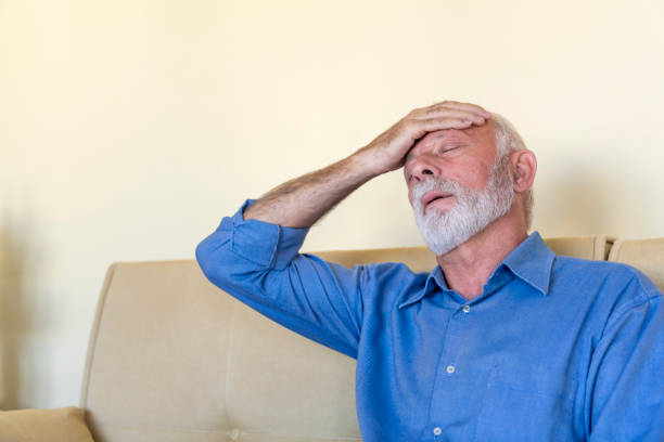 Photo of an Elderly Man Sitting on a Sofa in the Living Room at Home and Touching his Head Because of the Terrible Pain. Senior Man is Massaging his Painful Head.