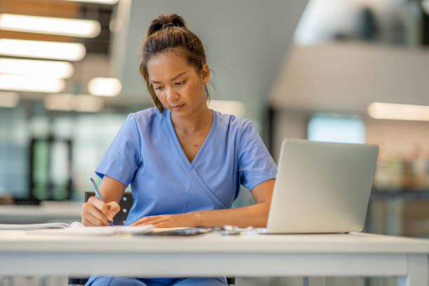 Medical student studying A woman wearing scrubs diligently studies at a desk in a modern educational building. medical student stock pictures, royalty-free photos & images