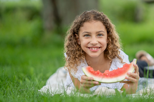 A girl lies on the grass in the summertime and eats a big slice of juicy watermelon.