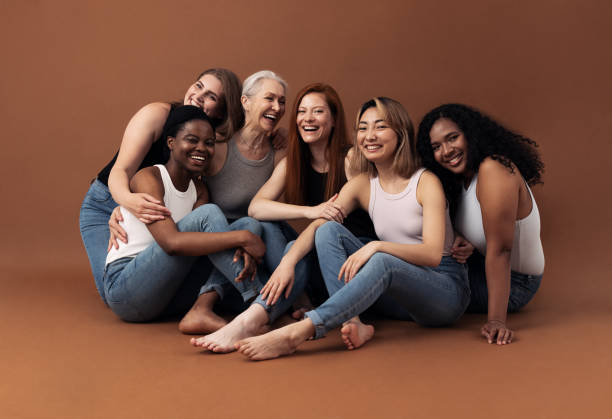 retrato de seis mujeres riendo de diferentes edades y tipos de cuerpo sentadas juntas sobre un fondo marrón en el estudio - diferencia entre generaciones fotografías e imágenes de stock