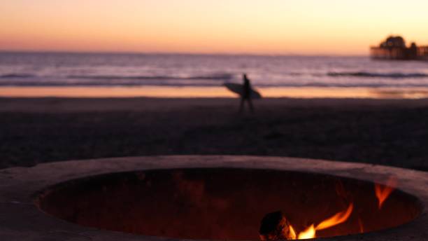 istock Campfire pit in California USA. Camp fire on twilight ocean beach, bonfire flame, surfer and waves. 1331893792