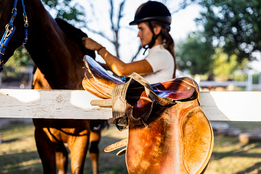 Beautiful and dedicated young woman in riding gear taking  care of her horse.She groomes her horse after riding, removes his saddle and other equipment.