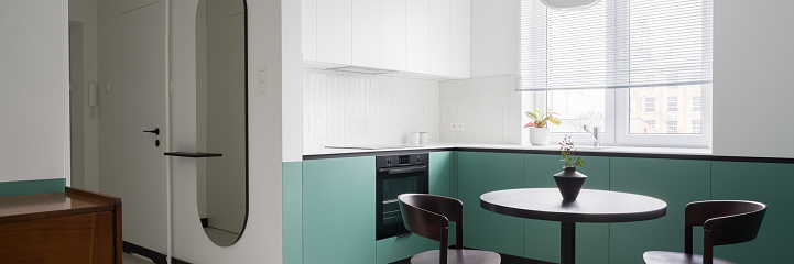 Panorama of stylish apartment with modern white and mint green kitchen cupboards with black dining table and chairs open to corridor with trendy oval mirror