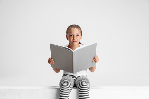 Fairy tail. Portrait of cute Caucasian girl, teen sitting and reading big book, magazine isolated on gray studio background. Happy childhood, education, emotion, facial expression concept.