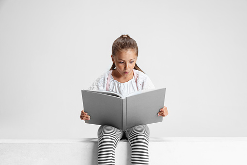 Portrait of cute Caucasian girl, teen sitting and reading big book, magazine isolated on gray studio background. Happy childhood, education, emotion, facial expression concept. Looks surprised