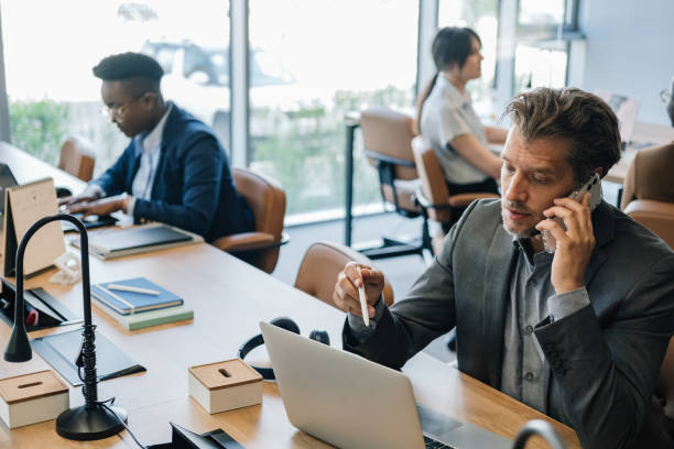 Displeased Business Man Talking on a Mobile Phone at Work Worried businessman arguing on the phone and working on laptop computer while sitting with team of mixed race colleagues and working together in an open plan office space. hot desking stock pictures, royalty-free photos & images