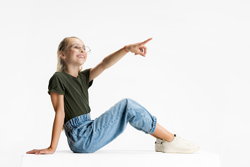 Pointing upwards. Side view portrait of one teen cute caucasian girl in casual clothes sitting on floor isolated over white studio background. Copyspace for ad.