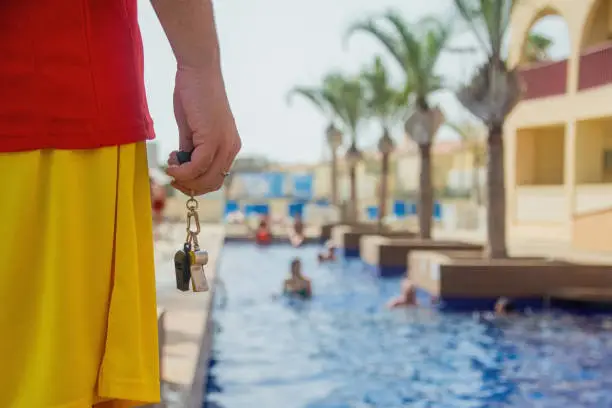 Close-up of a lifeguard's hand holding a whistle and watching over a swimming pool - Lifeguard at work concept