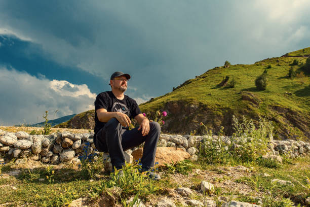 homme assis parmi les collines verdoyantes et le ciel nuageux seul - desaturated photos et images de collection
