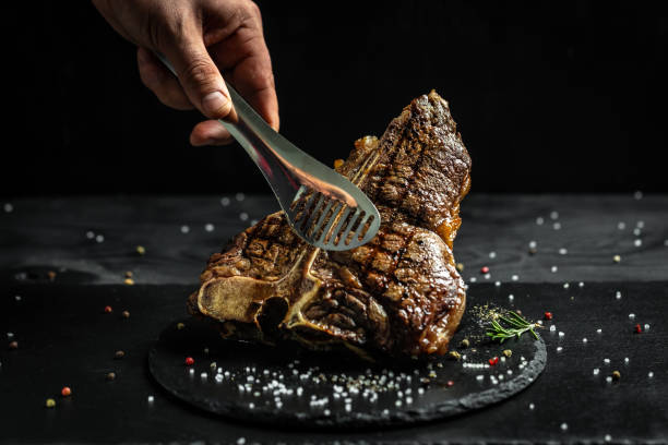chef hand holding steak meat tongs. large piece of fresh beef meat prepared on a grill. Medium rare Grilled T-Bone Steak, Barbecue aged wagyu porterhouse chef hand holding steak meat tongs. large piece of fresh beef meat prepared on a grill. Medium rare Grilled T-Bone Steak, Barbecue aged wagyu porterhouse. fire alphabet letter t stock pictures, royalty-free photos & images