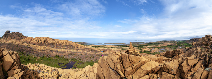 A wide panorama of the orange cliffs on the shore of the blue sea under the blue sky. Footpath to the observation view point. Rocks with serpentine minerals, Leka, Norway. The island is known for these rocks. Warm colors of the sunlight. Norway's geological national monument.