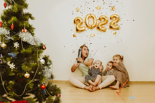 Mom and daughters with real emotions of joy explode a confetti clapper in the New Year 2022. Christmas family party at home with a Christmas tree and foil balls