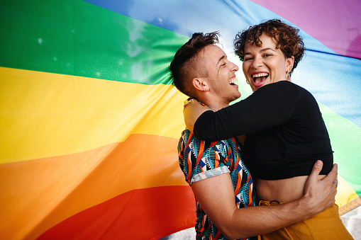 Happy queer couple standing against a rainbow pride flag. Young LGBTQ couple smiling cheerfully while embracing each other. Two non-conforming lovers celebrating gay pride together.