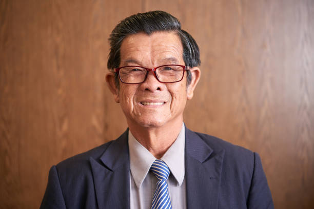Cheerful Senior Entrepreneur Portrait of cheerful senior entrepreneur in glasses standing against wooden wall and smiling at camera vietnamese old man portrait stock pictures, royalty-free photos & images