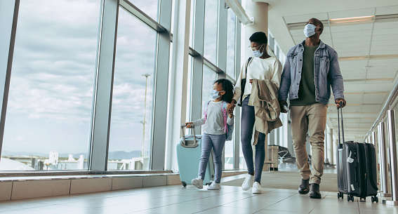 African family with luggage walking by window at airport terminal and watching airplanes through glass window. Family walking through airport passageway.