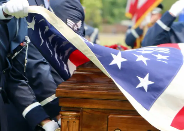 Selective focus white gloved hands of US Military Honor Guardsmen folding American flag over casket. Blurred faceless soldier in background.