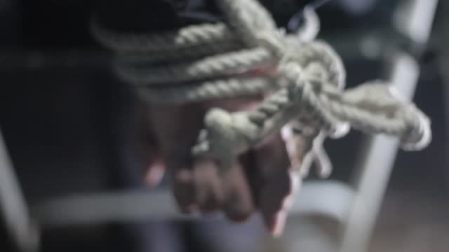 Close up of human hands tied with white colored rope and sitting on a metal chair.