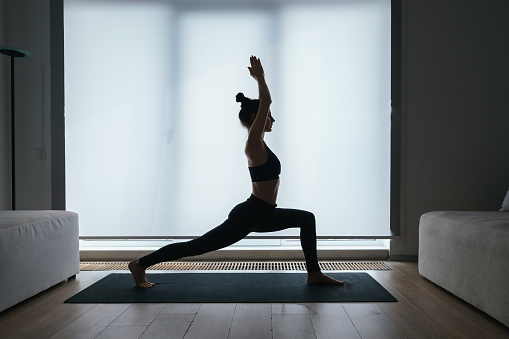 Silhouette of beautiful slim woman performing high crescent lunge yoga position while exercising at home, feeling calm and relaxed. Balance, zen and meditation concept