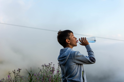 the boy drinks spring water from a plastic bottle, mountain background and clouds. A child drinks water from a bottle while walking, health. Boy drinks water from a bottle in clouds.
