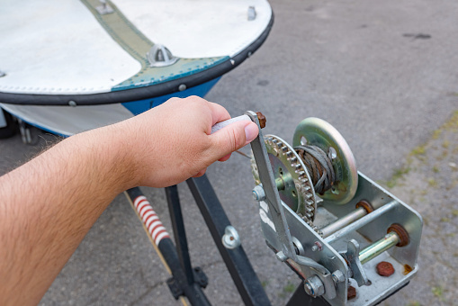 Person's hand turns the lever handle of the crank boat trailer winch. Preparing to travel and fishing.