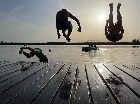 Silhouette of boys jumping. Horizon over water surface. Sky is clear and sun is shining. Phot of three boys jumping from the wooden dock caught in the air, after taking off.