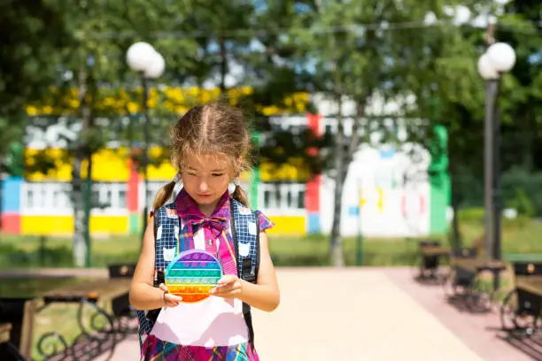Photo of Little girl with a backpack and in a school uniform in the school yard plays pop it toy. Back to school, September 1. The pupil relaxes after lessons. Primary education, elementary class for student