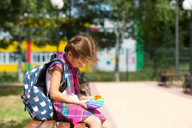 Photo of Little girl with a backpack and in a school uniform in the school yard plays pop it toy. Back to school, September 1. The pupil relaxes after lessons. Primary education, elementary class for student