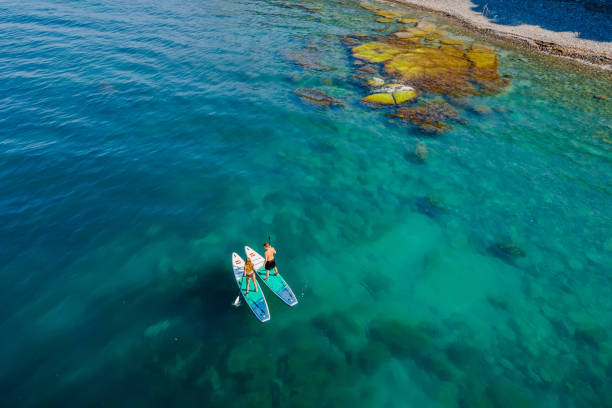 paar auf stand up paddle board am blauen meer mit felsen. menschen auf red paddle sup board in transparentem meer. luftbild - editorial women paddleboard surfboard stock-fotos und bilder