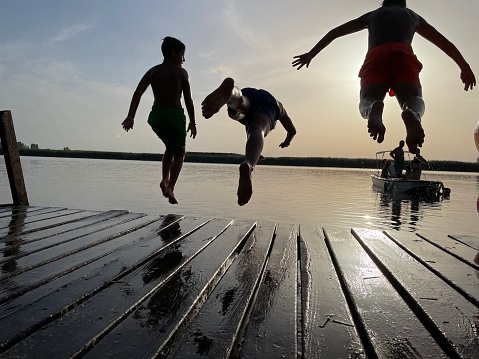 Three boys are jumping in the river from wooden jetty. Sunbeam. Silhouette of a boys jumping. Sky is clear and sun is shining. Horizon over water surface.